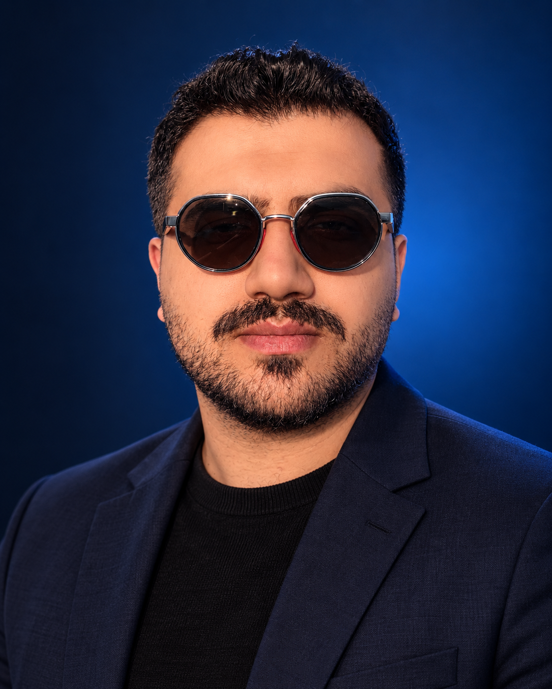 Ali Azmoudeh wearing a navy blazer and sunglasses in a studio-style portrait